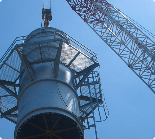 A large, cylindrical metal structure being lifted by a crane against a blue sky.