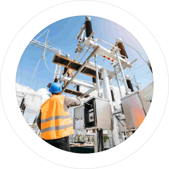 An engineer in a safety vest and helmet works on electrical equipment at a substation, surrounded by high-voltage power lines and clear skies.