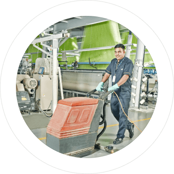 A worker operating an industrial floor cleaning machine in a factory setting, wearing a uniform and gloves.