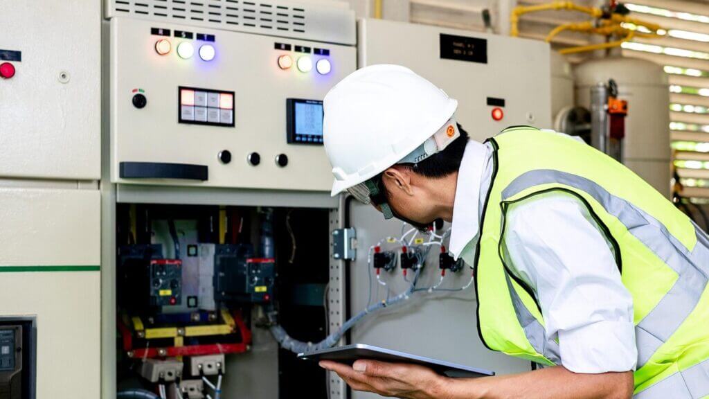 An engineer wearing a hard hat and safety vest inspects control panel equipment while holding a tablet.